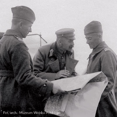Józef Piłsudski wyjaśnia coś oficerowi, trzyma w ręku obszerną płachtę papieru, zapewne mapę, oraz papierosa