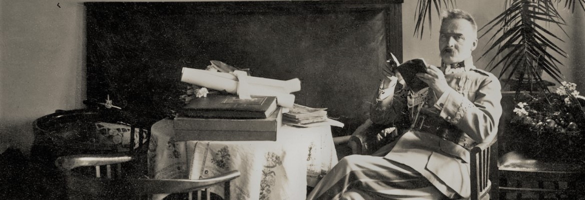 Józef Piłsudski w mundurze siedzi na krześle w salonie dworku Milusin trzymając książkę w ręku, od której odwrócił wzrok, aby spojrzeć w obiektyw aparatu