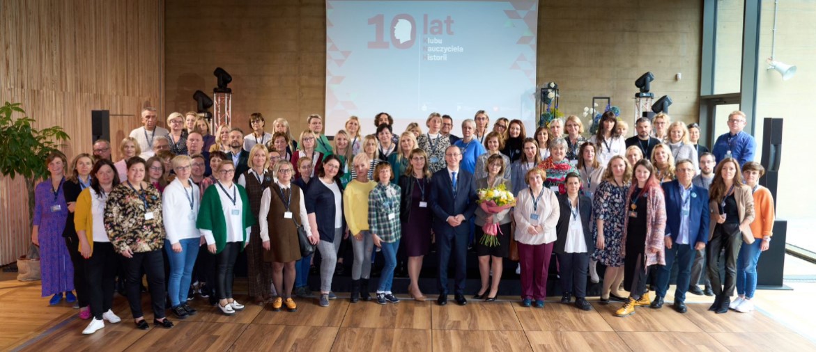 Zdjęcie przedstawia uczestniczki i uczestników X Zjazdu Klubu Nauczyciela Historii stojących w dużej, liczącej kilkadziesiąt osób grupie, zwróconych twarzami do aparatu (pozujących do zdjęcia). Zdjęcie zostało zrobione w Sali Paderewskiego w Muzeum Józefa Piłsudskiego w Sulejówku.