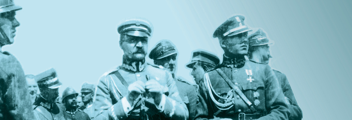 Józef Piłsudski w mundurze marszałkowskim trzyma w rękach małą lornetkę. Towarzyszą mu inni oficerowie Wojska Polskiego.
